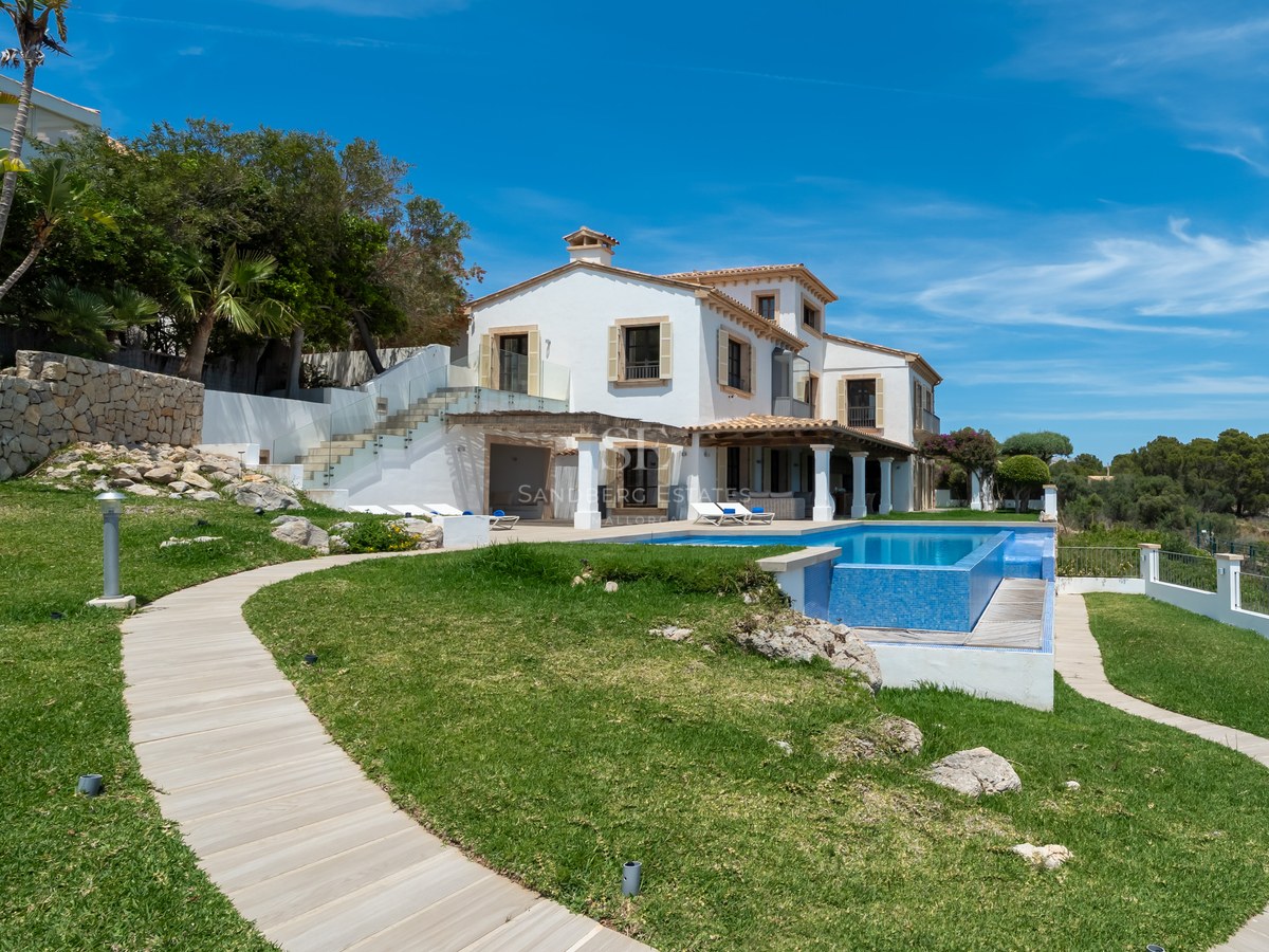 Villa méditerranéenne blanche avec piscine bleue, jardin paysager et chemin en bois sous un ciel bleu.