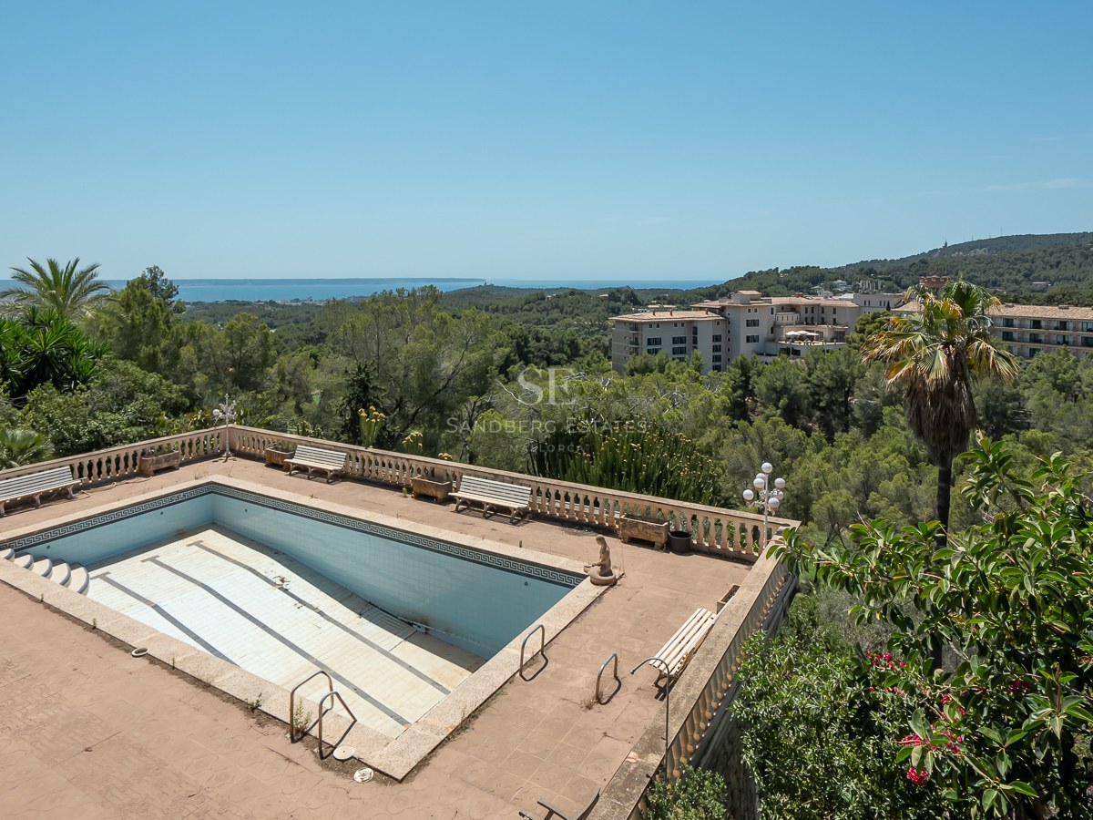 Piscina con balaustrada de piedra con vistas a un bosque frondoso y al mar Mediterráneo al fondo bajo un cielo despejado.