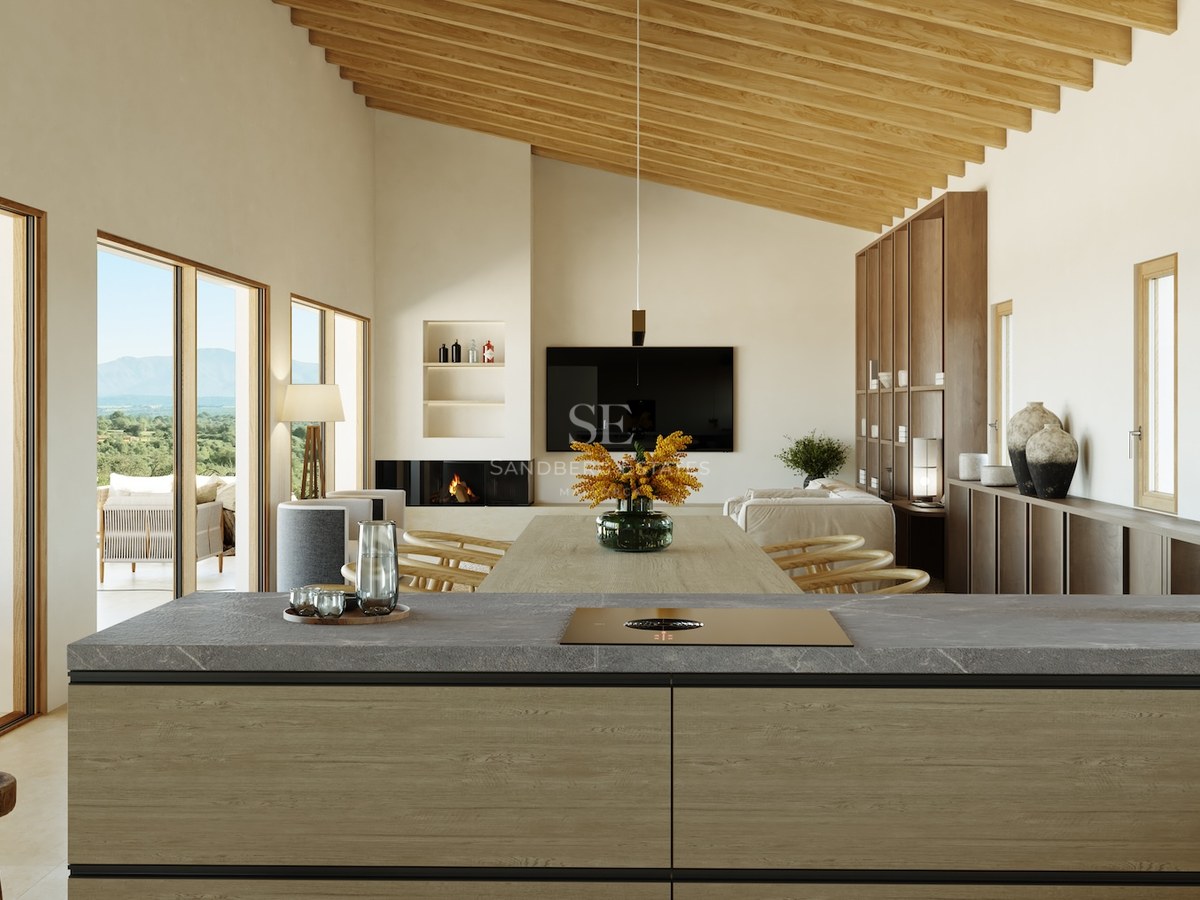 Innenansicht von der Kücheninsel zum Wohnzimmer mit Holzbalken, Kamin und großen Fenstern mit Landschaftsblick.