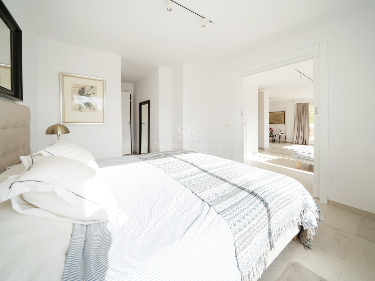 Dormitorio principal luminoso con paredes blancas, suelos cerámicos grandes, cabecero acolchado y vistas al salón.