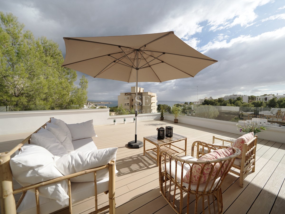Terraza en azotea con suelo de madera, muebles de bambú, sombrilla beige y barandillas de cristal frente al mar.
