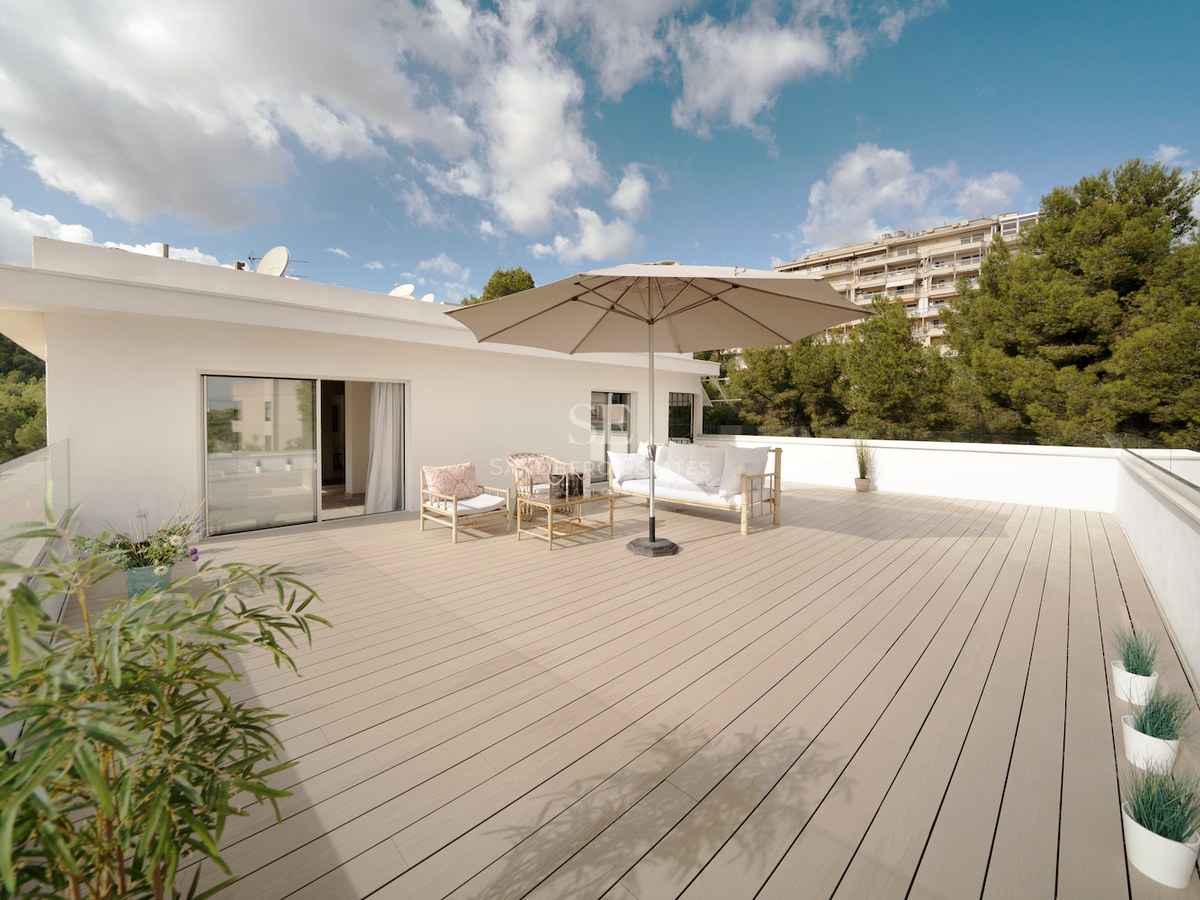 Terraza espaciosa con tarima de madera, muebles de exterior blancos y barandillas de cristal bajo un cielo despejado.