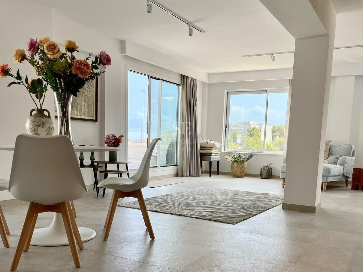 Salón amplio con comedor blanco, alfombra con texturas y grandes ventanales con vistas a la costa y jardines.