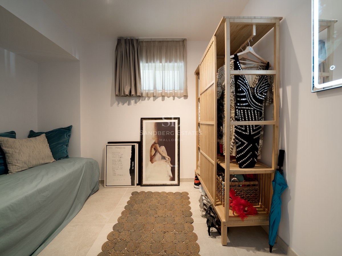 Vestidor moderno con armario de bambú abierto, un diván azul verdoso y cuadros apoyados en el suelo de baldosas.