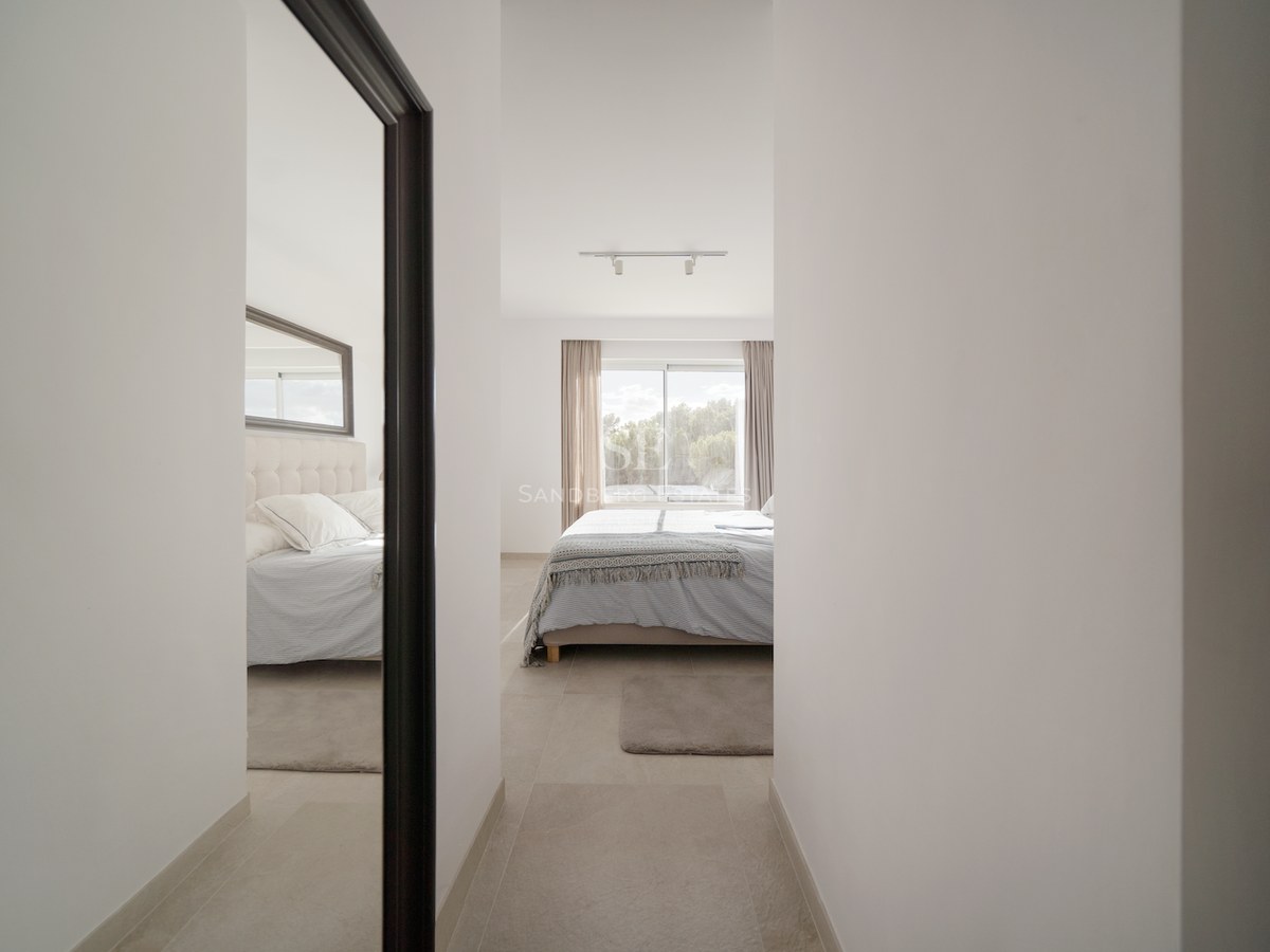 Vista desde un pasillo hacia un dormitorio luminoso con ventanal, textiles neutros y un espejo de cuerpo entero.