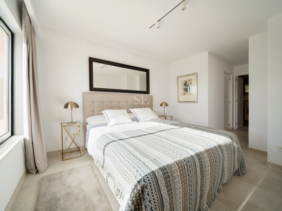 Dormitorio moderno con cama doble, cabecero tapizado, lámparas de latón y un gran espejo bajo luz natural.