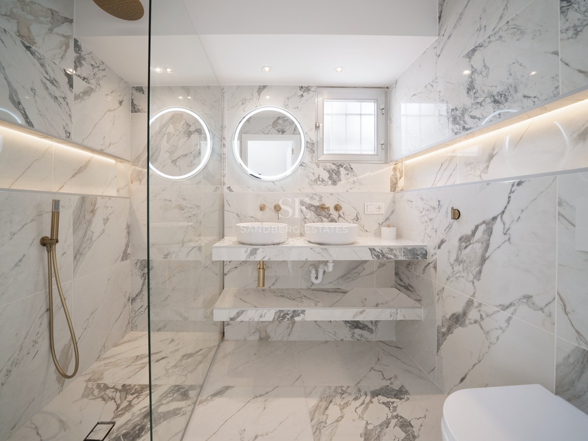 Baño de lujo con paredes de mármol, lavabo doble, espejos circulares retroiluminados y grifería de latón cepillado.