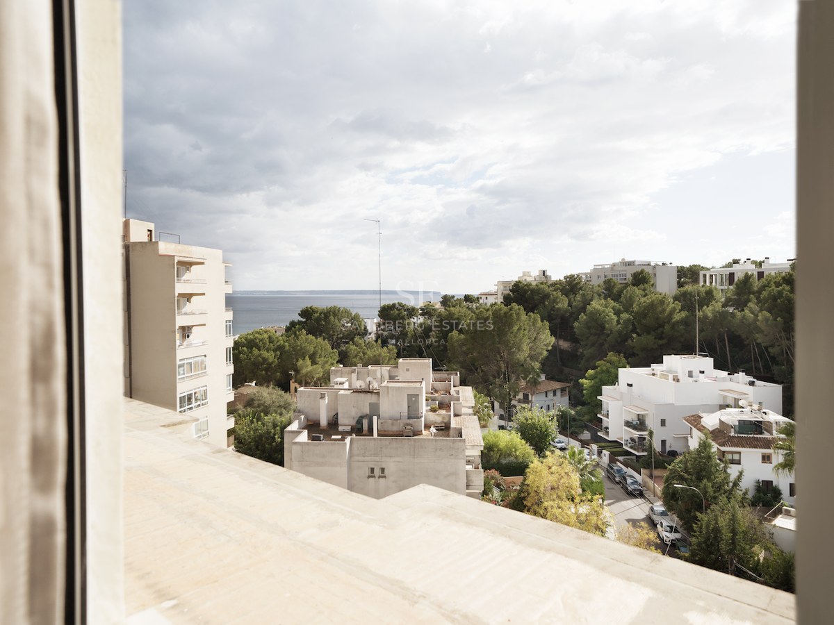 Vista elevada desde una ventana hacia edificios blancos, pinos y el horizonte del mar bajo un cielo nublado.