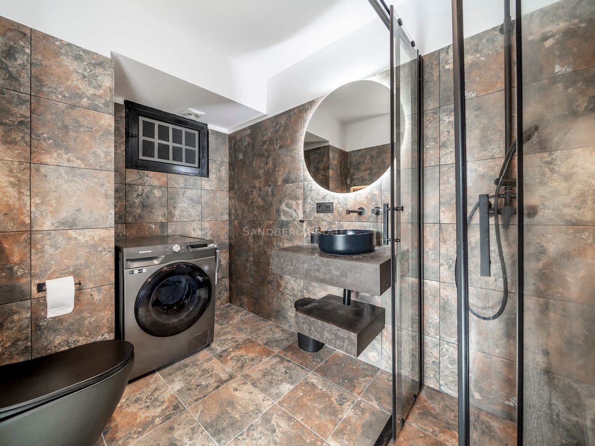 Baño contemporáneo con azulejos efecto piedra, lavabo suspendido negro, espejo retroiluminado y ducha de cristal.