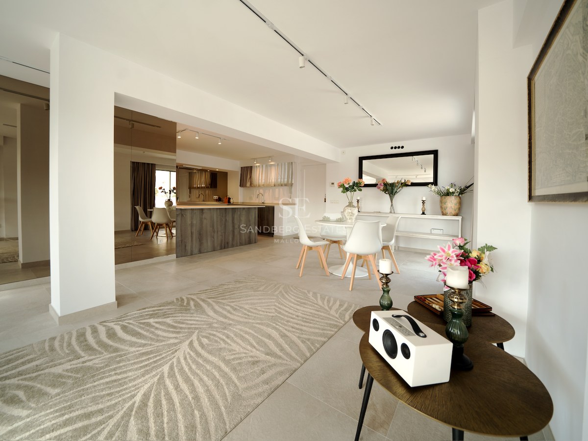 Espacio interior luminoso con comedor blanco, isla de madera, espejos de pared y suelos de gres porcelánico claro.
