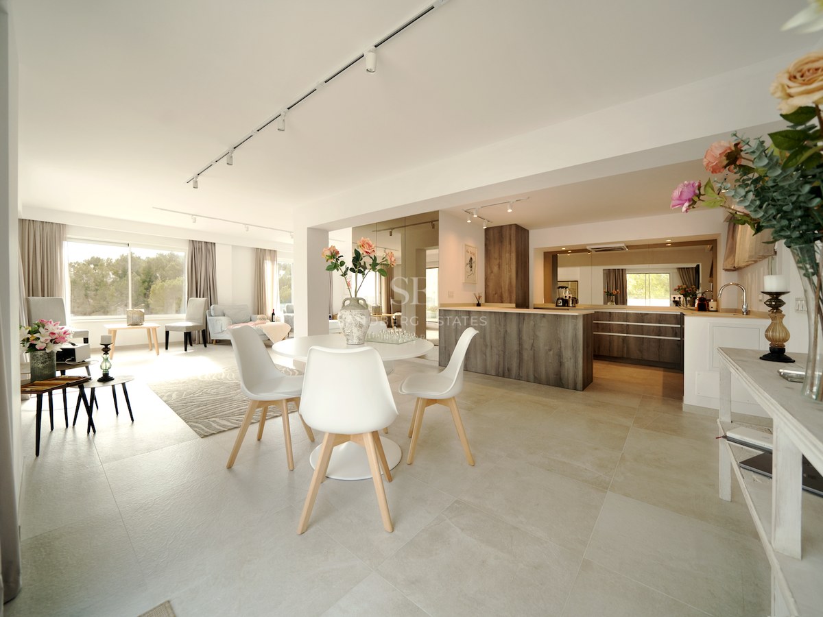 Amplia sala de planta abierta con mesa de comedor blanca, suelos de porcelánico y cocina con acabados en madera.