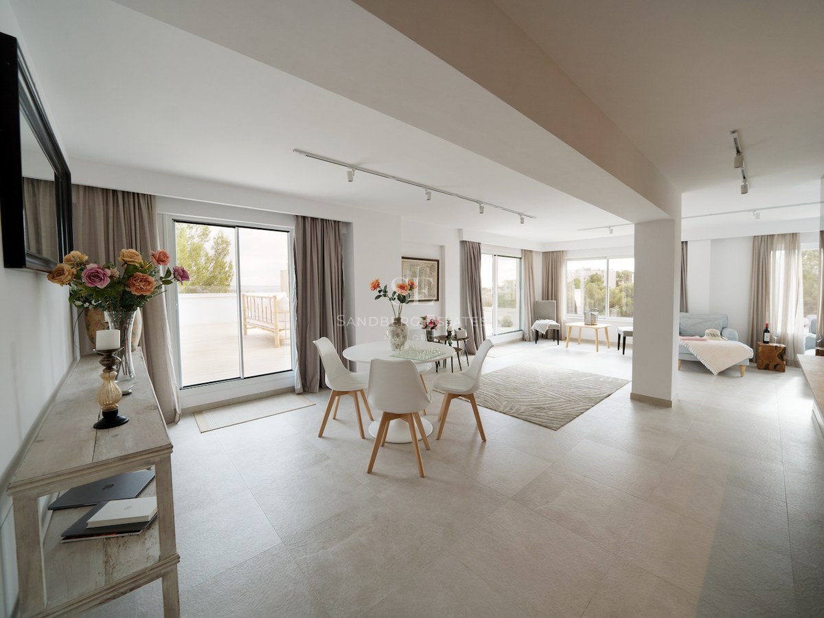 Comedor moderno con muebles blancos, suelos de porcelánico claro y grandes ventanales que dan a una terraza exterior.