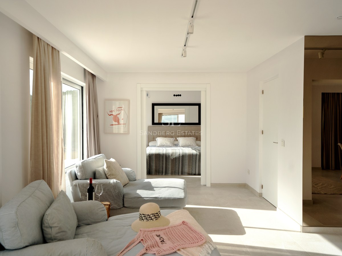 Suite de dormitorio moderna y luminosa con sofás azul claro, cortinas de lino y cama visible al fondo.