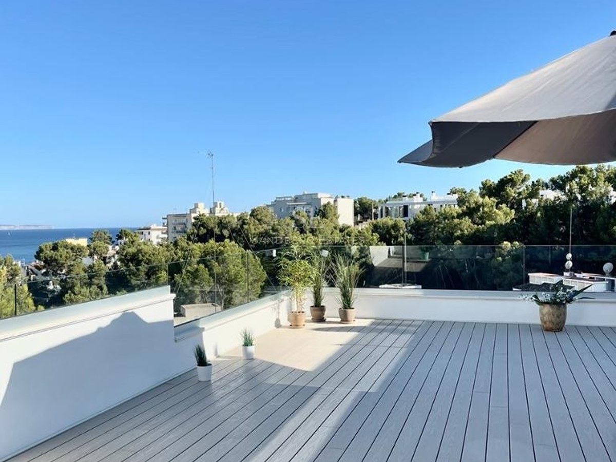 Terraza moderna con suelo de madera gris, barandillas de cristal y vistas panorámicas al mar y vegetación mediterránea.