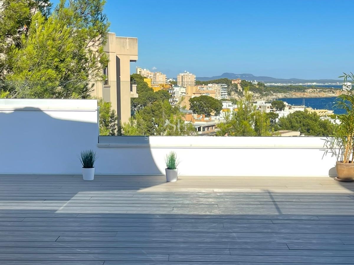 Terraza con suelo de madera gris y barandillas de cristal con vistas a la costa mediterránea y cielo azul despejado.