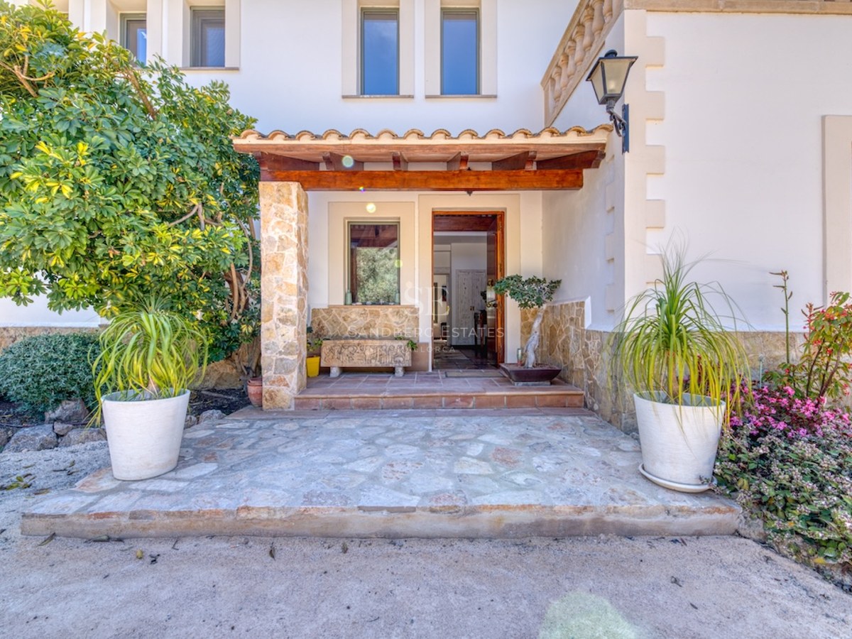 Entrée de villa avec piliers en pierre, porche en bois, murs blancs et plantes en pots sous une lumière naturelle.