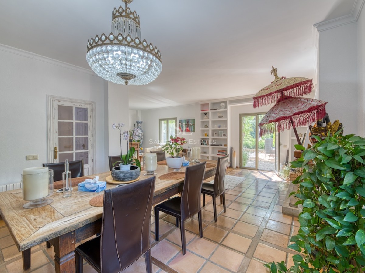 Salle à manger avec sol en terre cuite, table en bois rustique, chaises en cuir et lustre en cristal.
