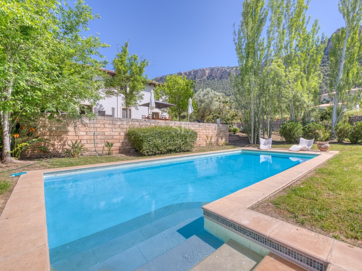 Piscine rectangulaire turquoise entourée d'une terrasse en pierre et d'un jardin avec des arbres et des montagnes.