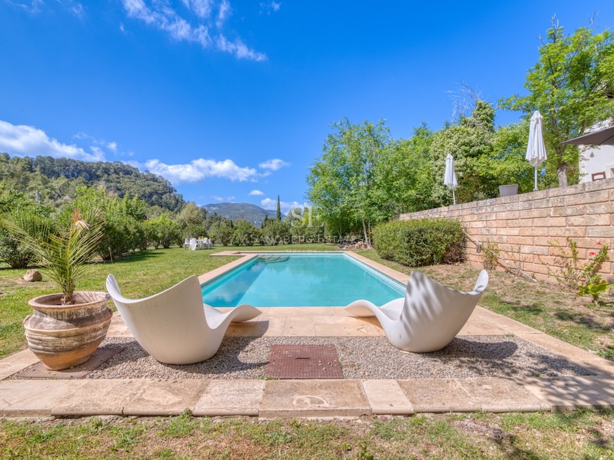 Piscine rectangulaire entourée d'une terrasse en pierre, pelouse verte, deux chaises longues blanches et vue montagne.