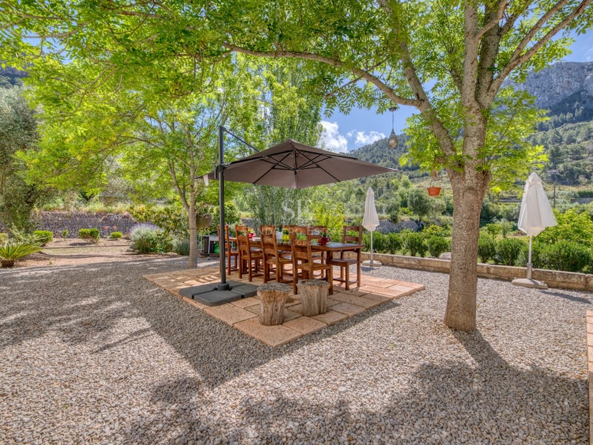 Table et chaises en bois sous un parasol sur une terrasse en gravier entourée d'arbres et de montagnes.