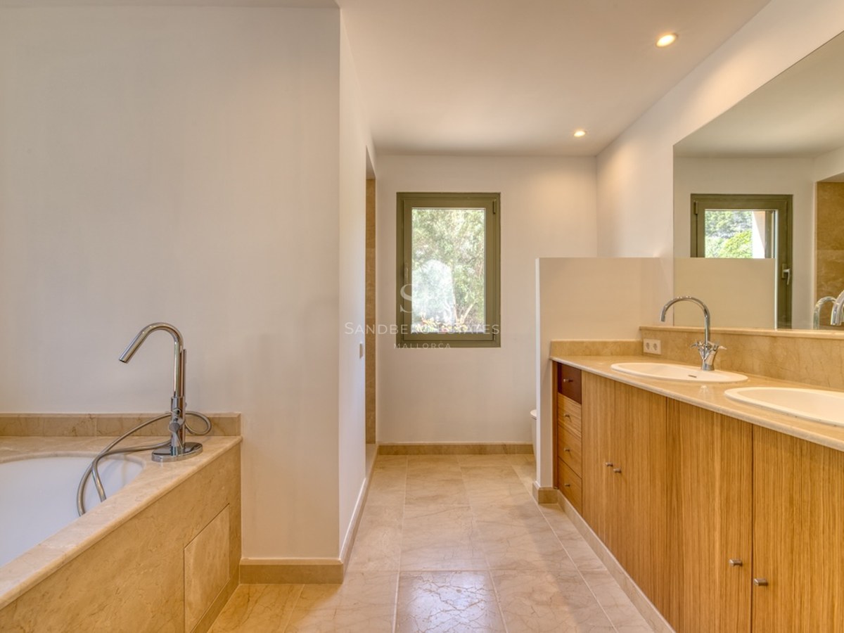 Salle de bain de luxe avec double vasque en bois, comptoirs en marbre beige, baignoire et lumière naturelle.