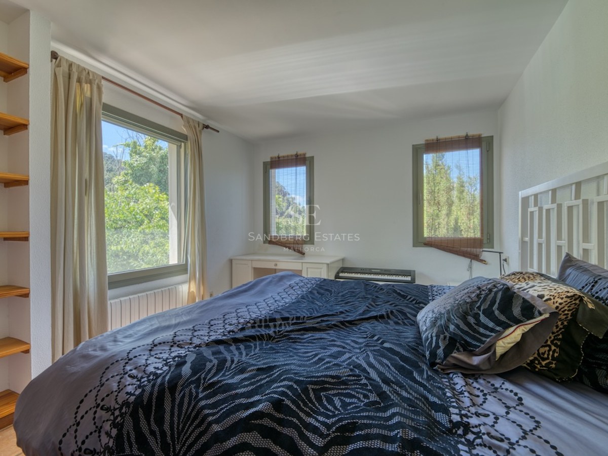 Chambre aux murs blancs, trois fenêtres donnant sur des arbres, lit avec coussins à motifs et étagères en bois.