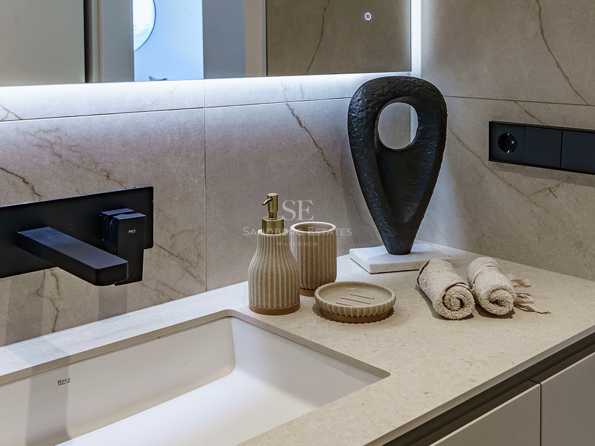 Närbild på ett vitt tvättställ med mattsvart kran, beige marmorvägg och designaccessoarer.