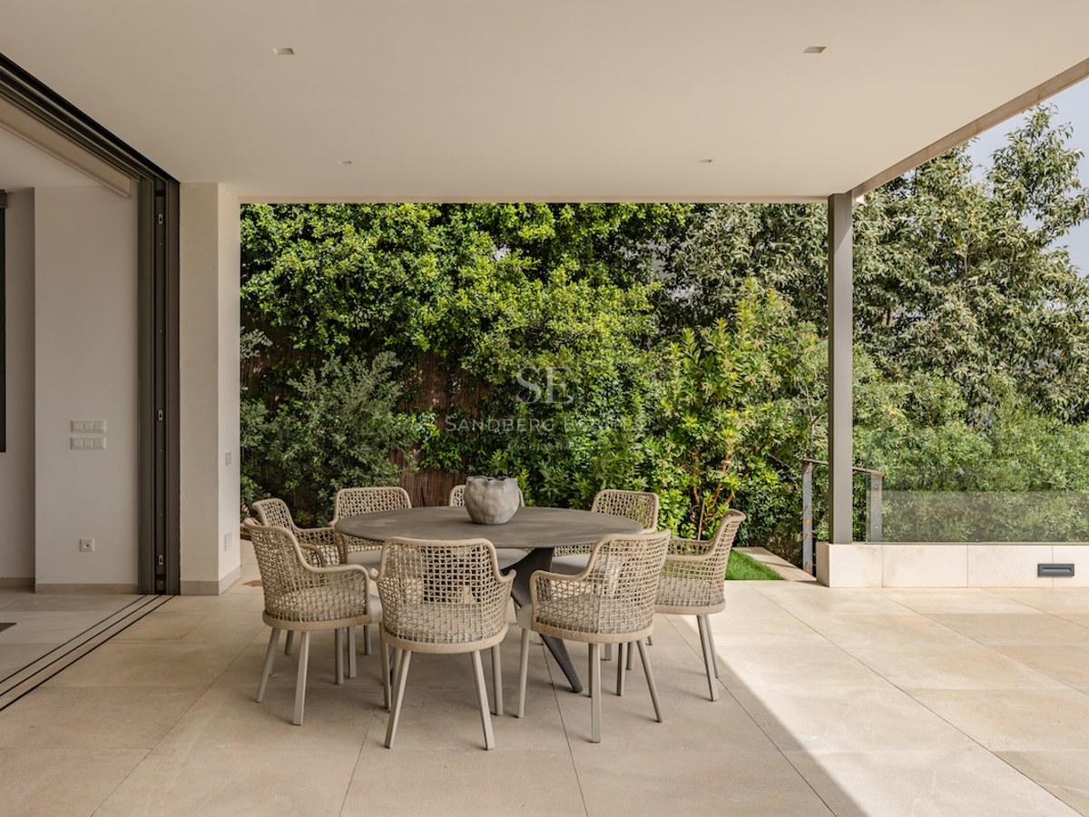 Modern covered terrace featuring a round dining table with woven chairs against a lush green garden backdrop.