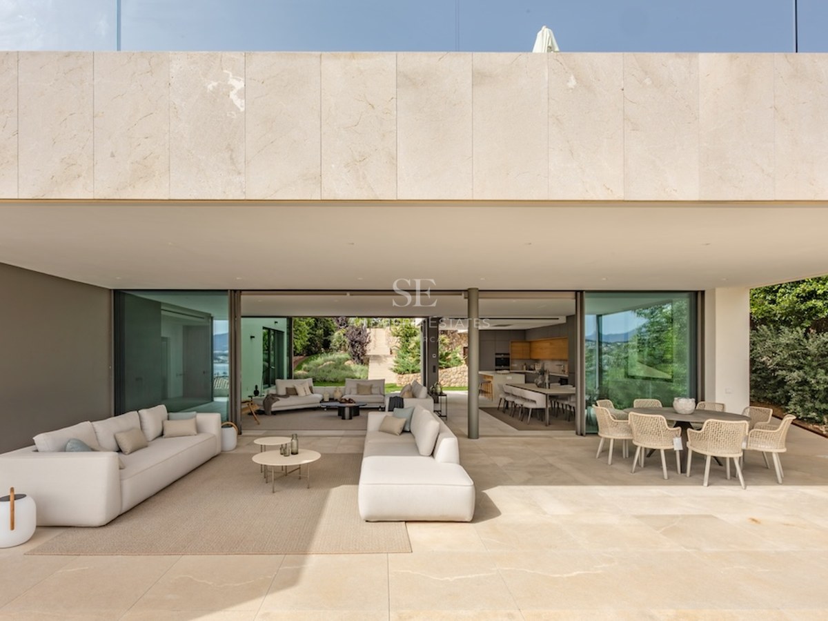 Luxury villa terrace featuring beige stone flooring, white sofas, and glass doors opening to living areas.