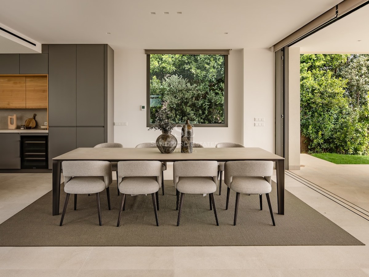 Modern dining room featuring a large rectangular table, eight beige upholstered chairs, and floor-to-ceiling glass doors.