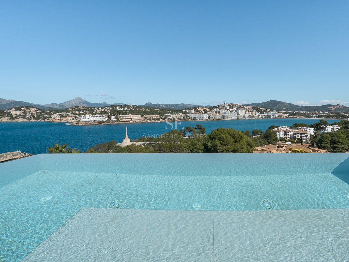 En infinitypool med utsikt över en blå vik, kustbyggnader och berg under en klar himmel.