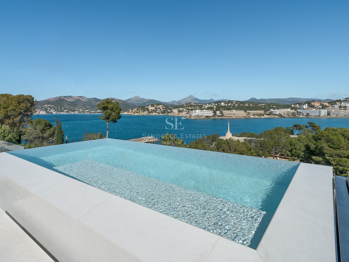 Modern infinitypool med turkost vatten som blickar ut över en blå vik, berg och kustlinje.