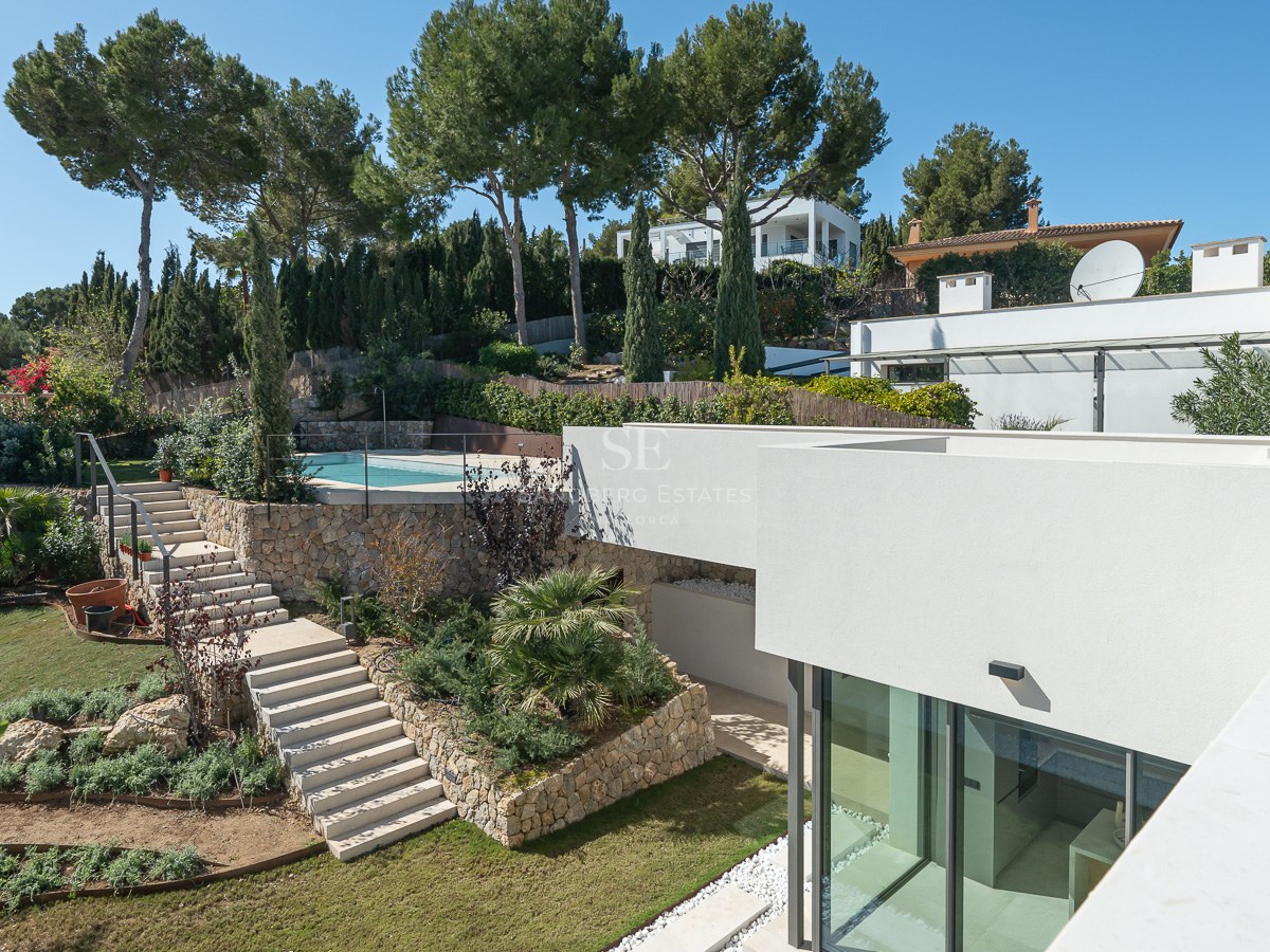 Modern vit villa med en terrasserad trädgård, stentrappor och en pool omgiven av tallar under en klarblå himmel.