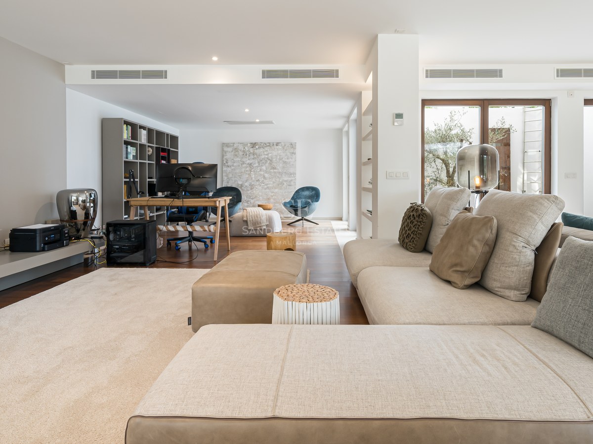 Salón moderno con sofá beige en forma de L, suelos de madera noble y zona de oficina al fondo con escritorio.