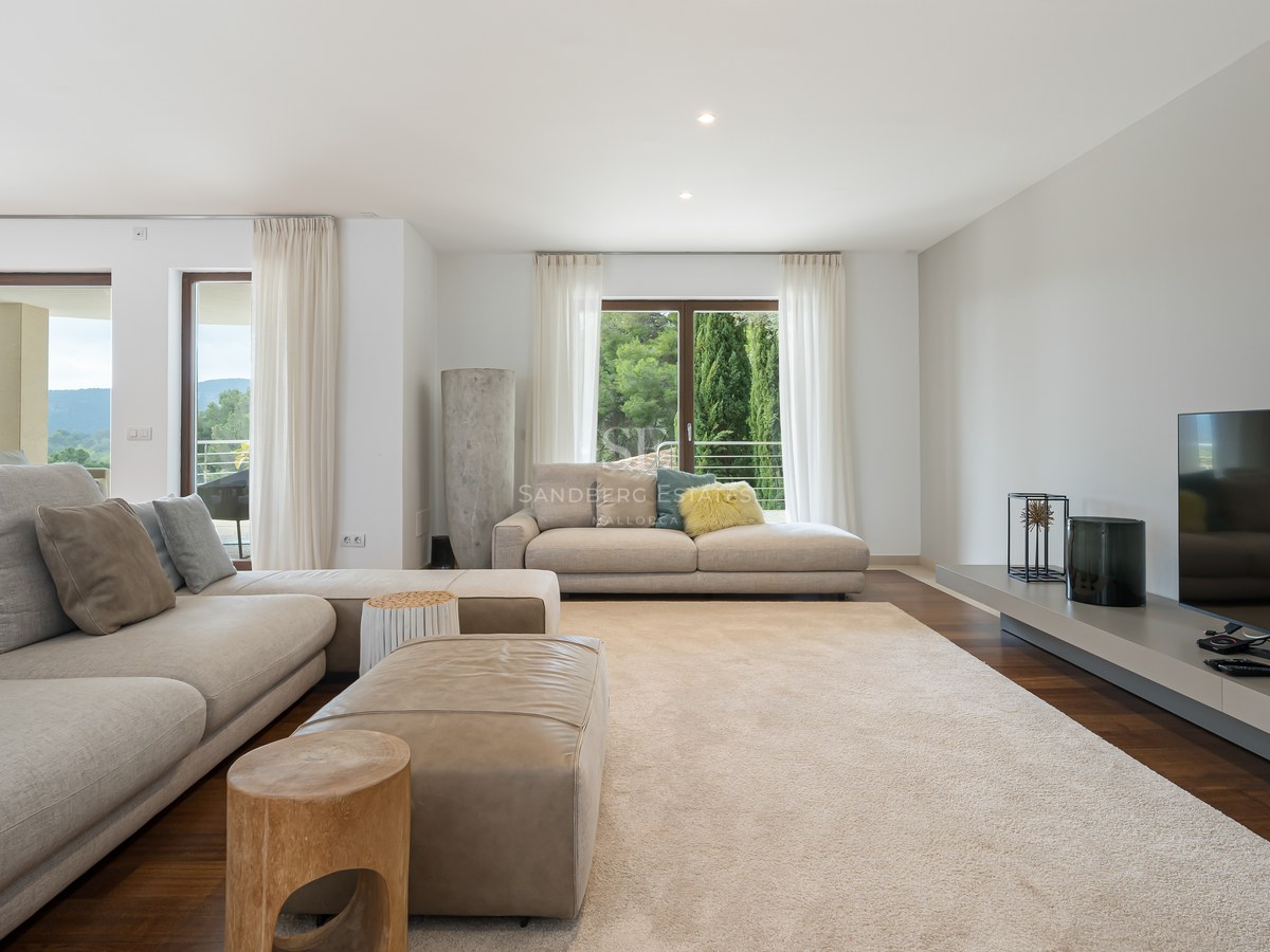 Amplio salón con sofás beige, otomana de cuero, suelos de madera oscura y ventanales con vistas a colinas verdes.