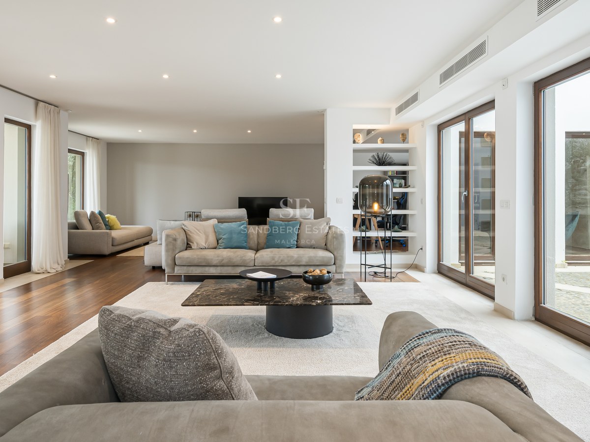 Amplio salón moderno con suelos de madera, sofás grises, mesa de centro de mármol y ventanales de suelo a techo.