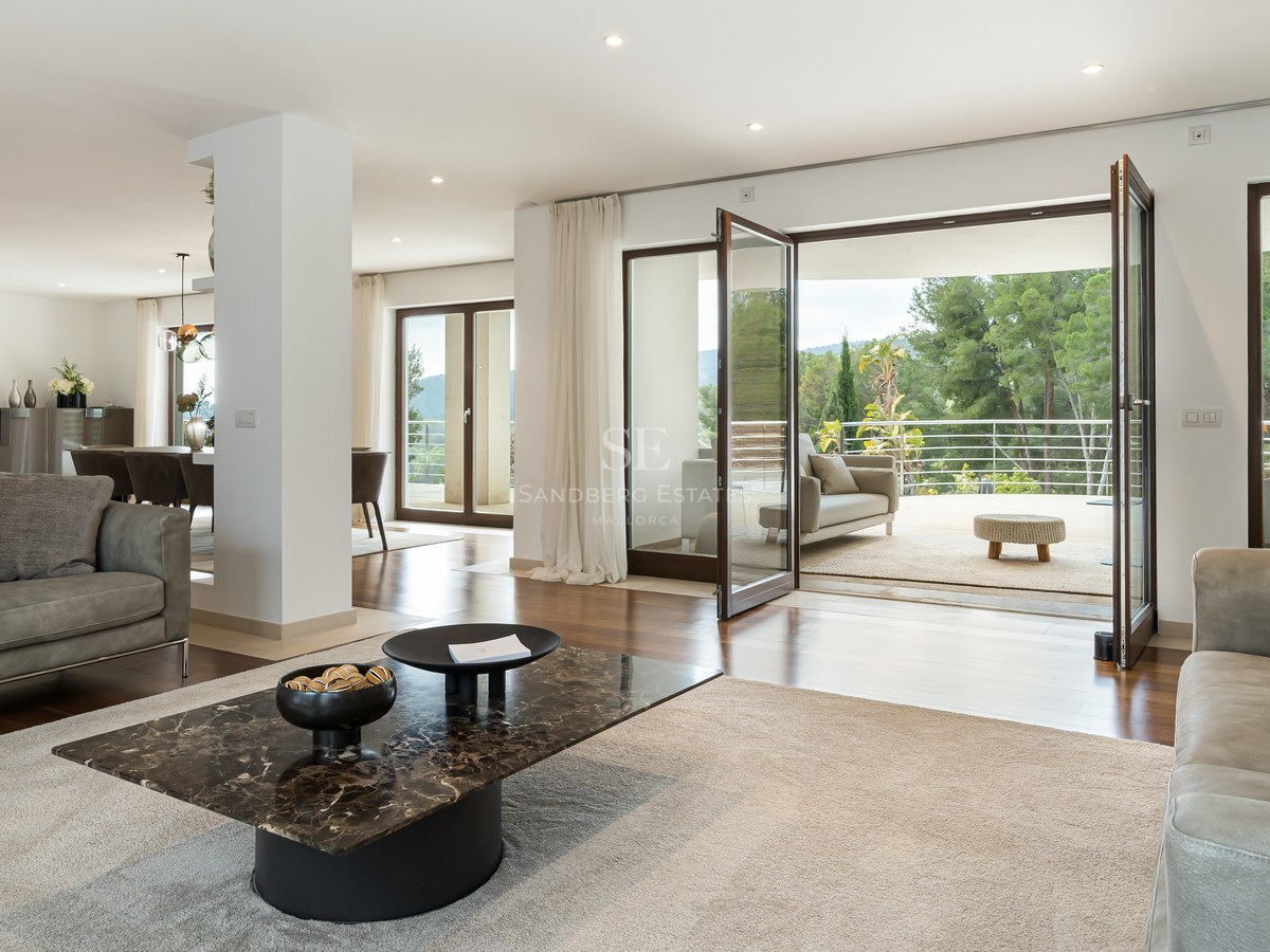 Salón moderno con sofás de cuero, mesa de mármol y grandes puertas de cristal que dan a una terraza con vistas al bosque.