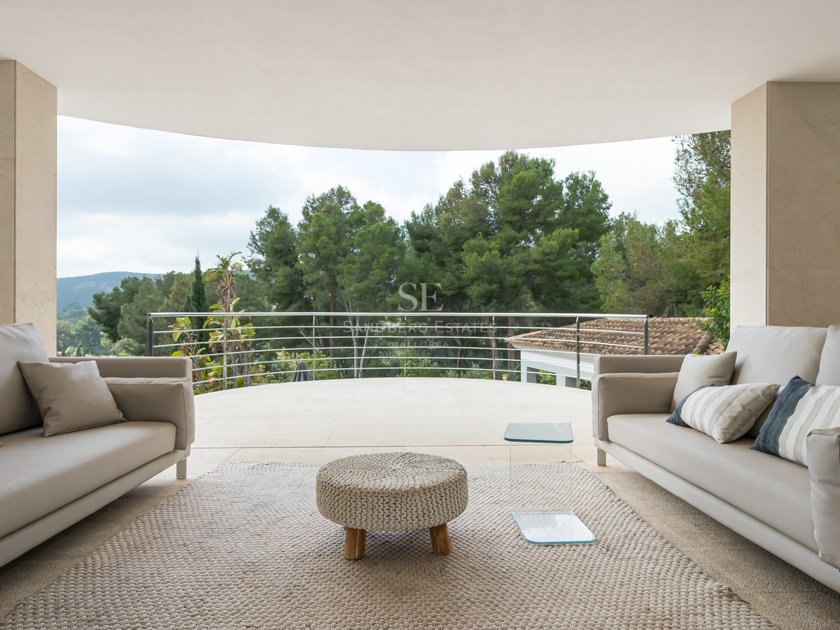 Terraza moderna con sofás grises y puf tejido frente a un bosque denso bajo un cielo despejado.