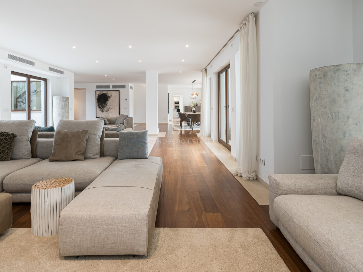 Amplio salón moderno con sofás de tela gris, suelos de madera oscura y grandes ventanales de cristal.