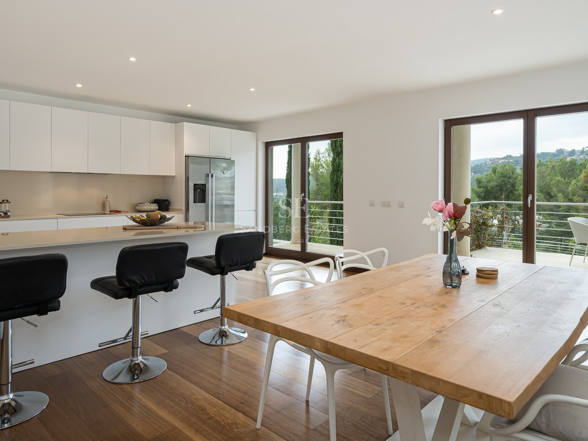 Cocina blanca contemporánea con barra americana, mesa de comedor de madera y ventanales con vistas al exterior.