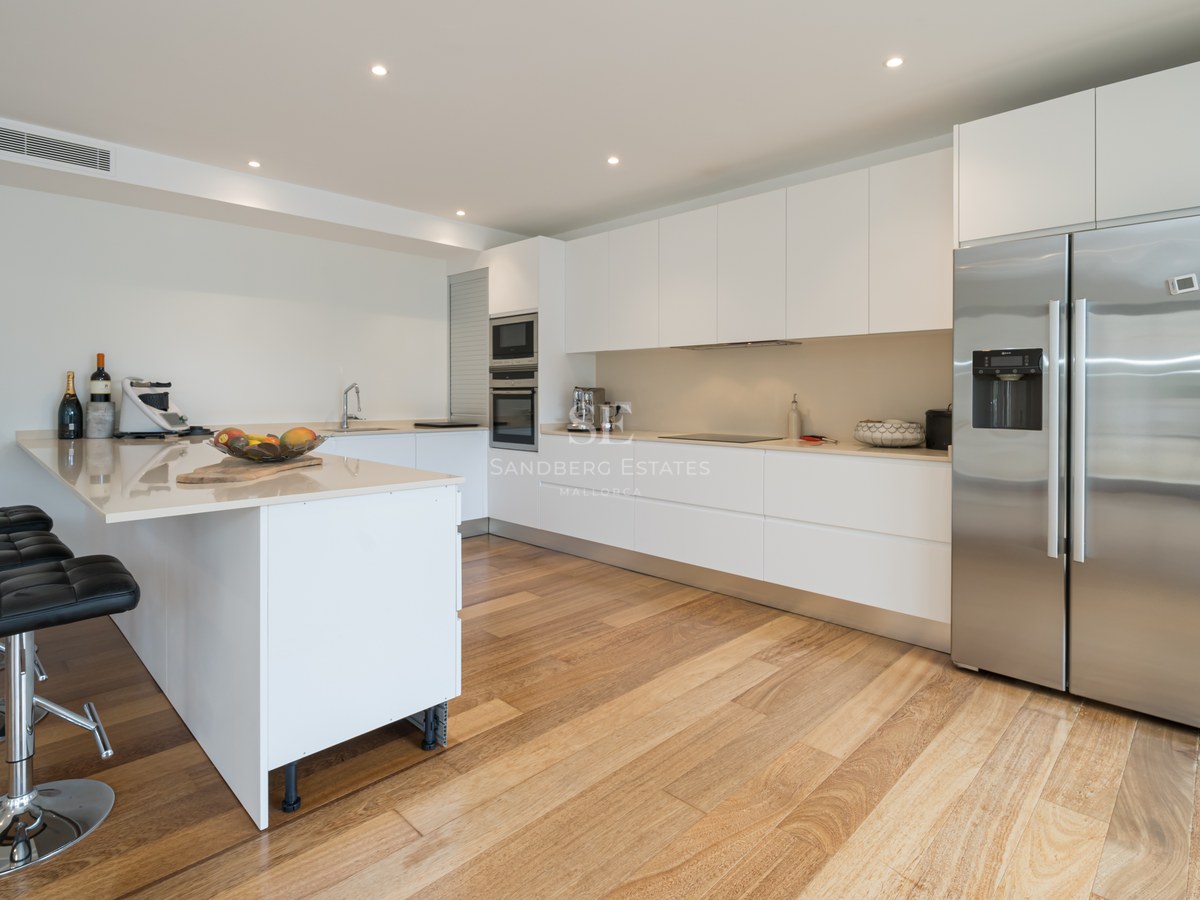 Cocina blanca con suelo de madera, frigorífico de acero inoxidable, encimera clara y taburetes de cuero negros.