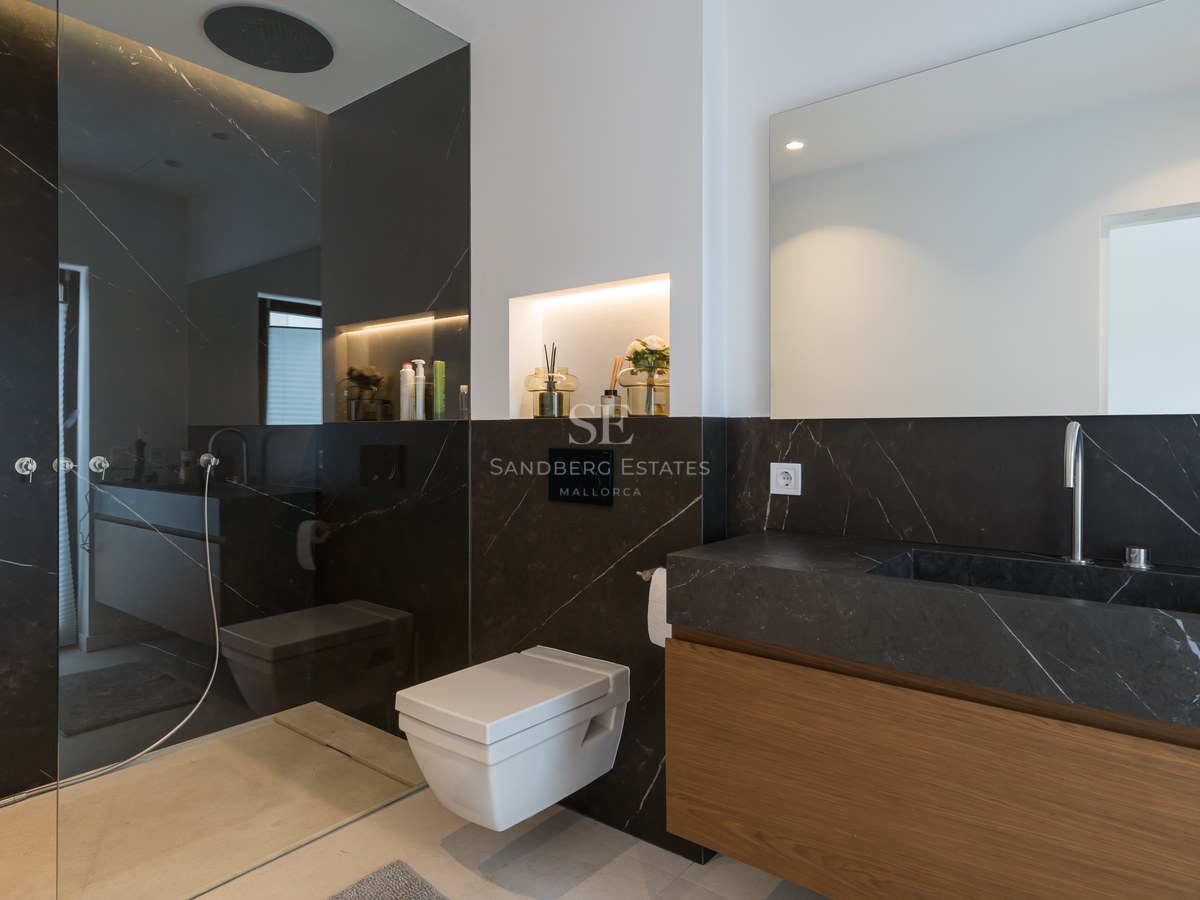 Baño moderno con paredes de mármol negro, inodoro suspendido, mueble de madera y ducha de obra acristalada.