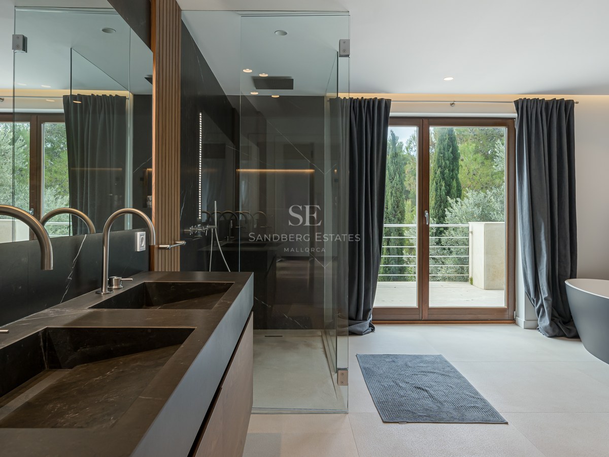 Baño moderno con lavabo doble negro, ducha de cristal, bañera exenta y acceso a terraza con vistas a la naturaleza.