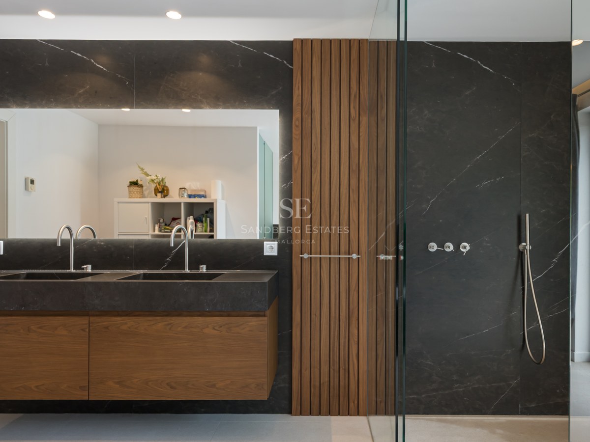 Baño principal moderno con lavabo doble de mármol negro, paneles de madera de nogal y amplia ducha de cristal.