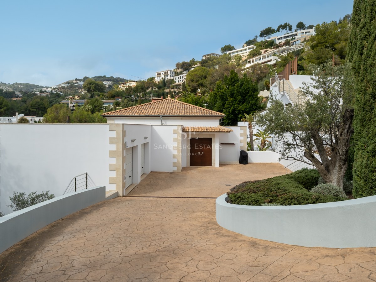 Entrada privada pavimentada hacia una villa blanca con tejado de terracota, garaje doble y vegetación mediterránea.
