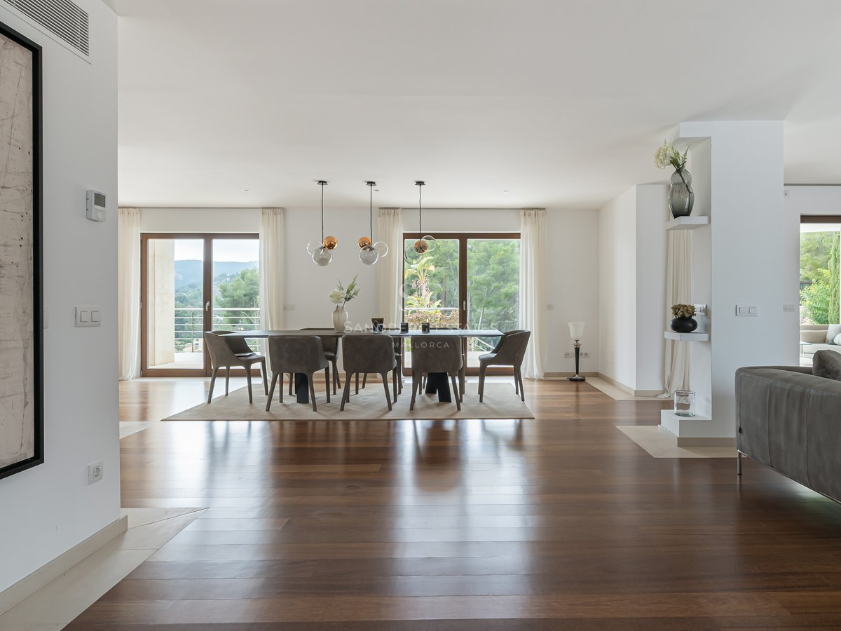 Comedor espacioso con suelos de madera oscura, lámparas de diseño y grandes ventanales hacia un jardín frondoso.