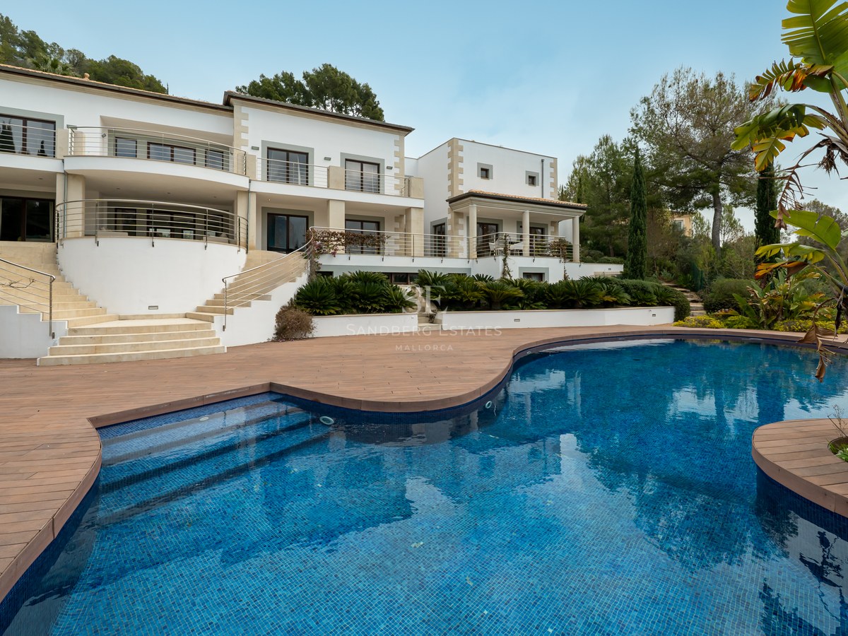Exterior de una villa blanca de lujo con una gran piscina curva y tarima de madera rodeada de vegetación.