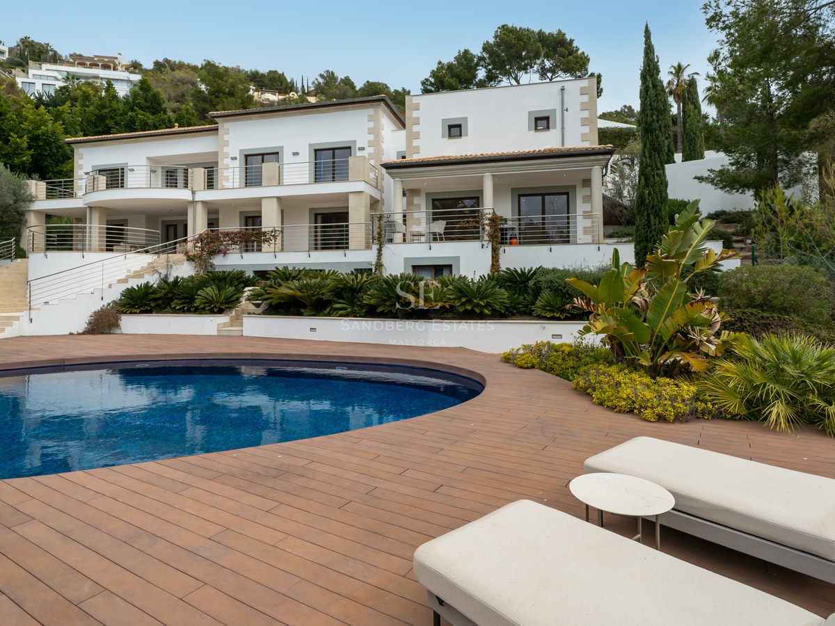 Villa de lujo blanca con piscina curva, amplia terraza de madera y tumbonas frente a una fachada moderna.