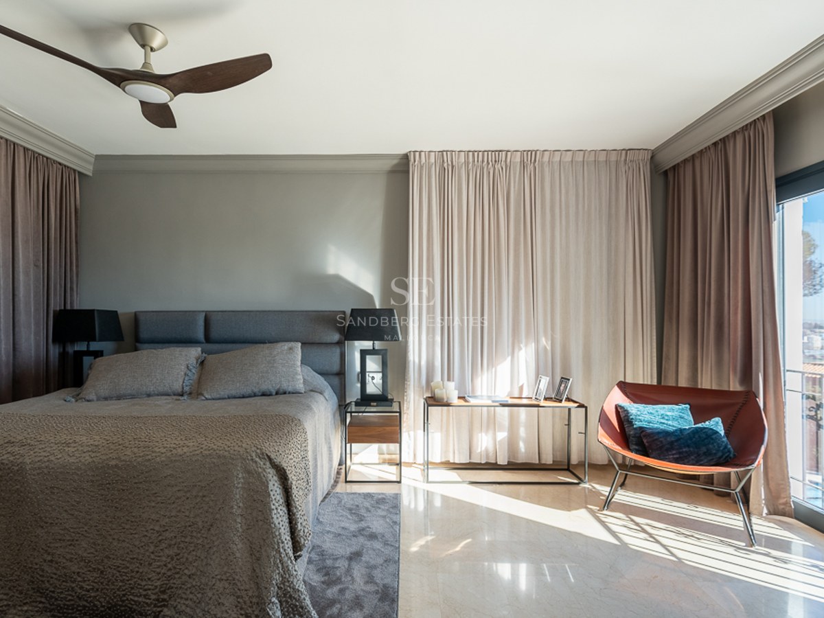 Dormitorio moderno con cama tapizada gris, suelo de mármol, sillón de cuero y balcón con vistas a la costa.