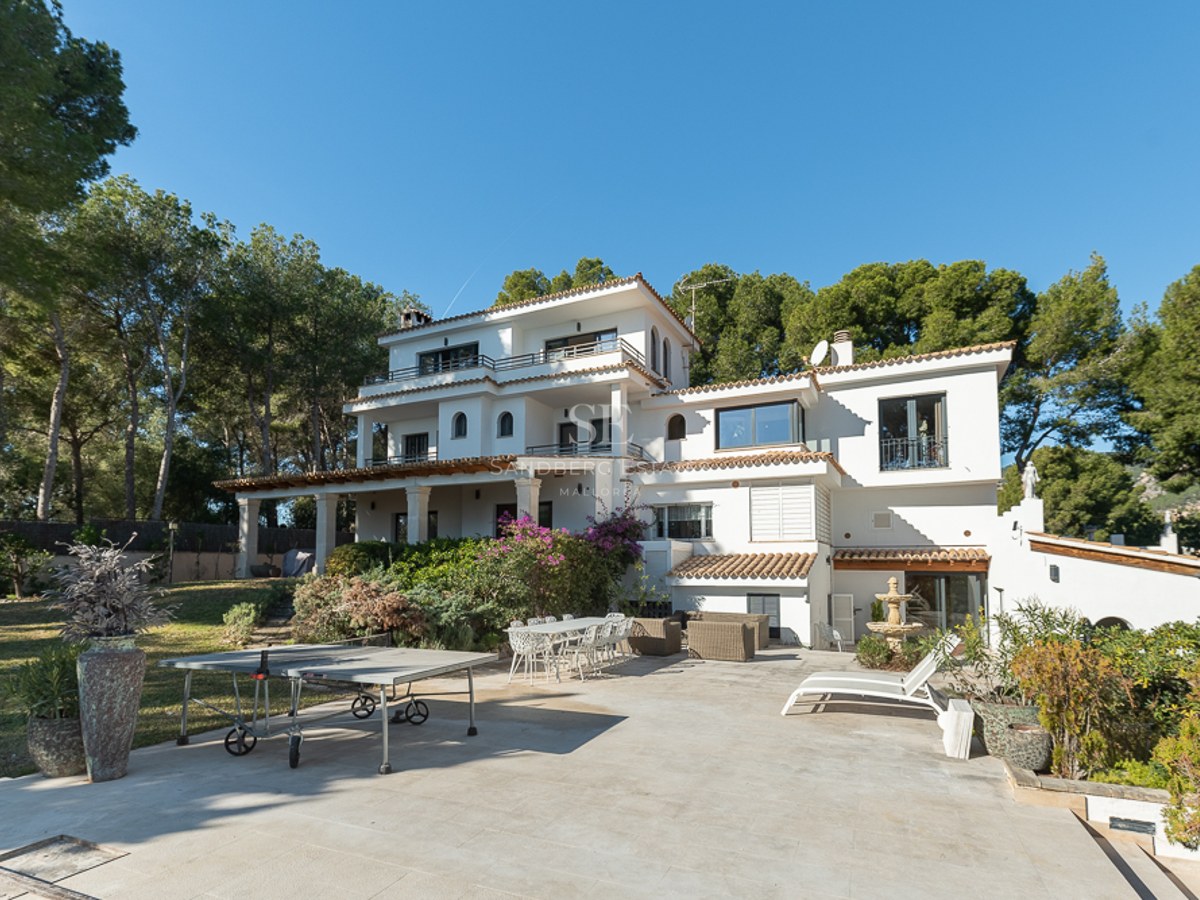 Vista exterior trasera de una villa mediterránea blanca de tres niveles con terrazas de piedra y entorno boscoso.
